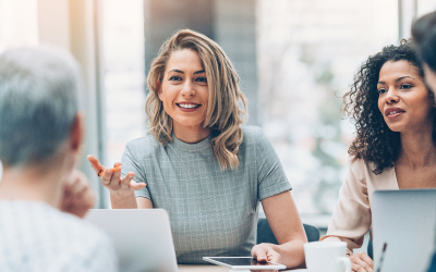 Group of professional women engaged in a friendly discussion during a business meeting, with one woman smiling and gesturing while holding a tablet.