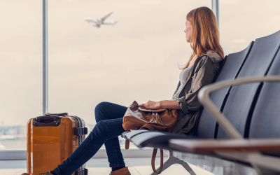 A person sits in an airport terminal beside a suitcase, looking out the window at an airplane taking off or landing.