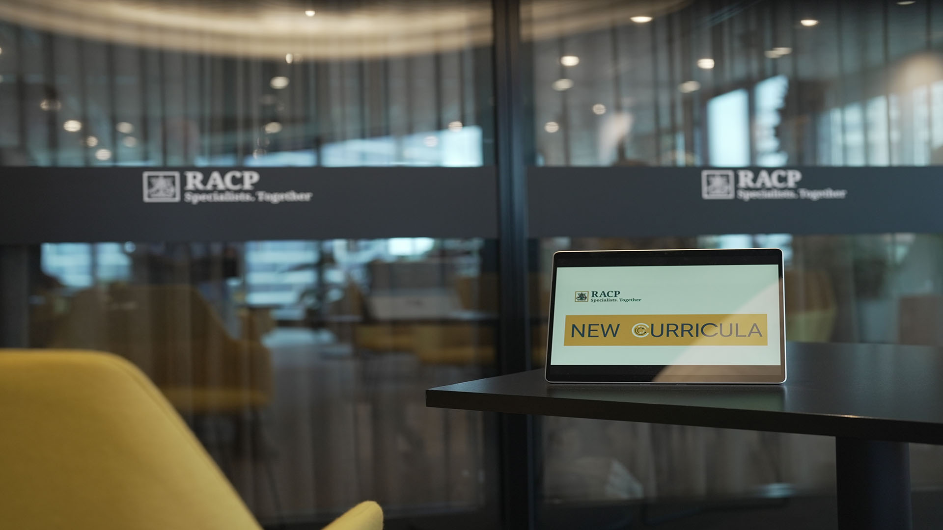 Image of a tablet on a desk in the RACP lobby with a screensaver image saying curricula renewal