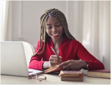 Woman writing down notes in book in front of laptop