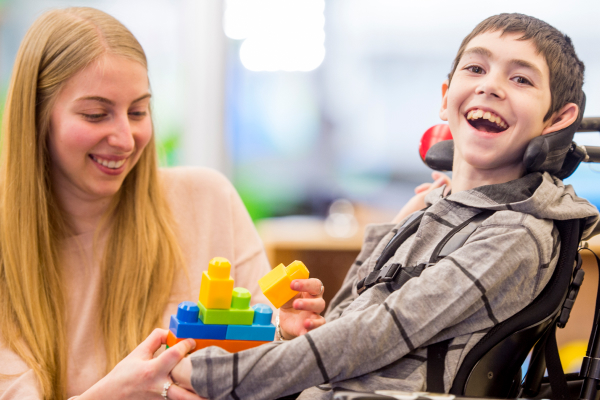 Smiling child in a wheelchair playing with colourful building blocks alongside a supportive adult.