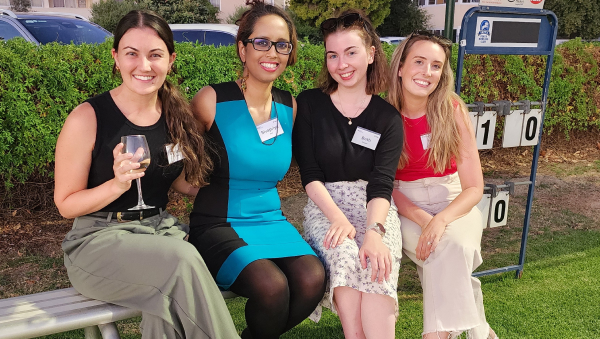 Four people sitting together on an outdoor bench at an event, dressed in semi-formal clothing, with a scoreboard and greenery in the background.