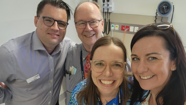 Four people standing together in a clinical room with medical equipment on the wall behind them/