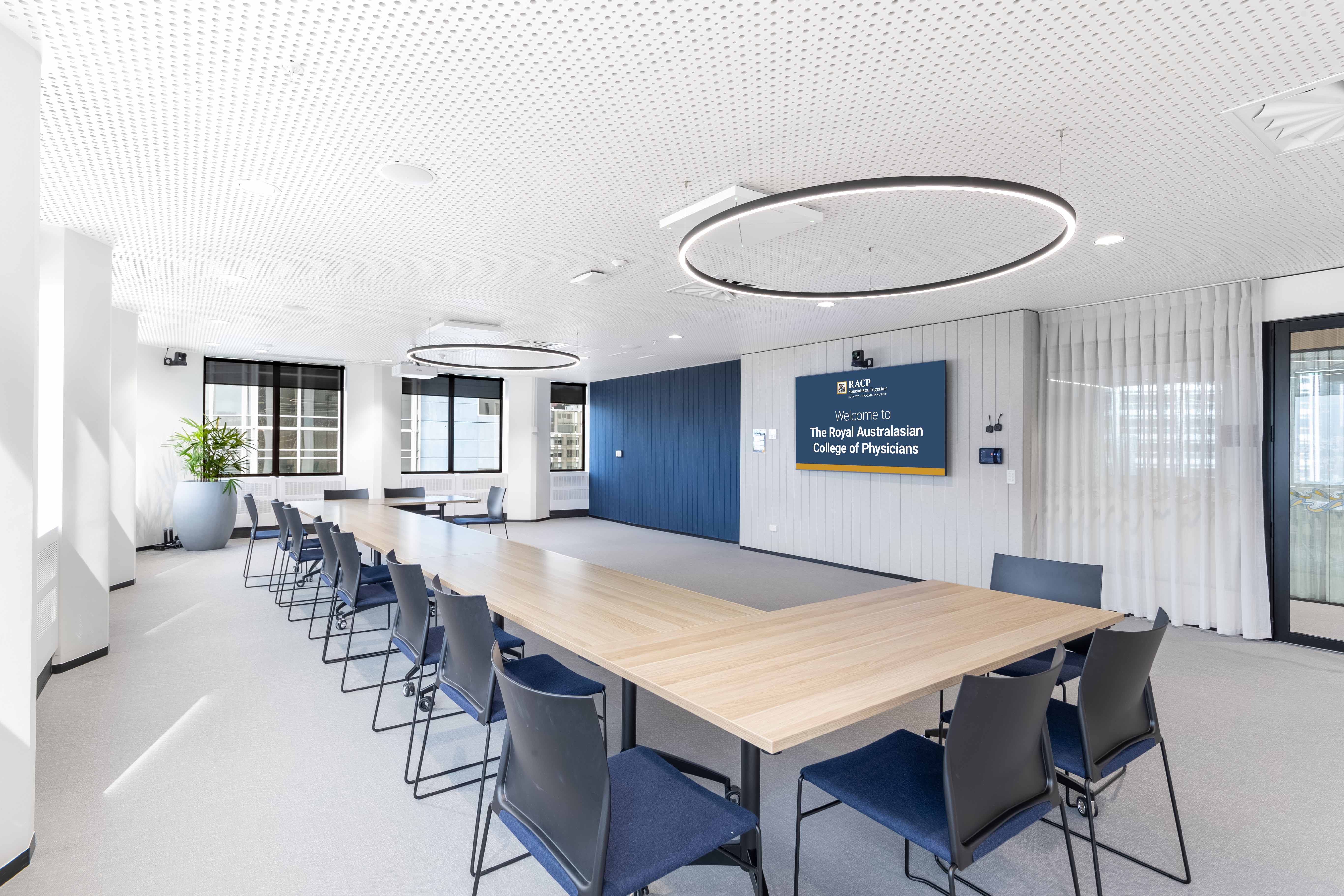 Modern meeting room with a long boardroom style table and chairs facing a presentation screen displaying a Royal Australasian College of Physicians welcome slide.