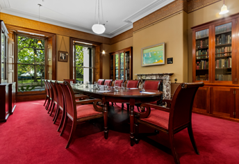 Traditional boardroom with a long polished table and red chairs, surrounded by wood paneling and large windows, set up for a meeting.