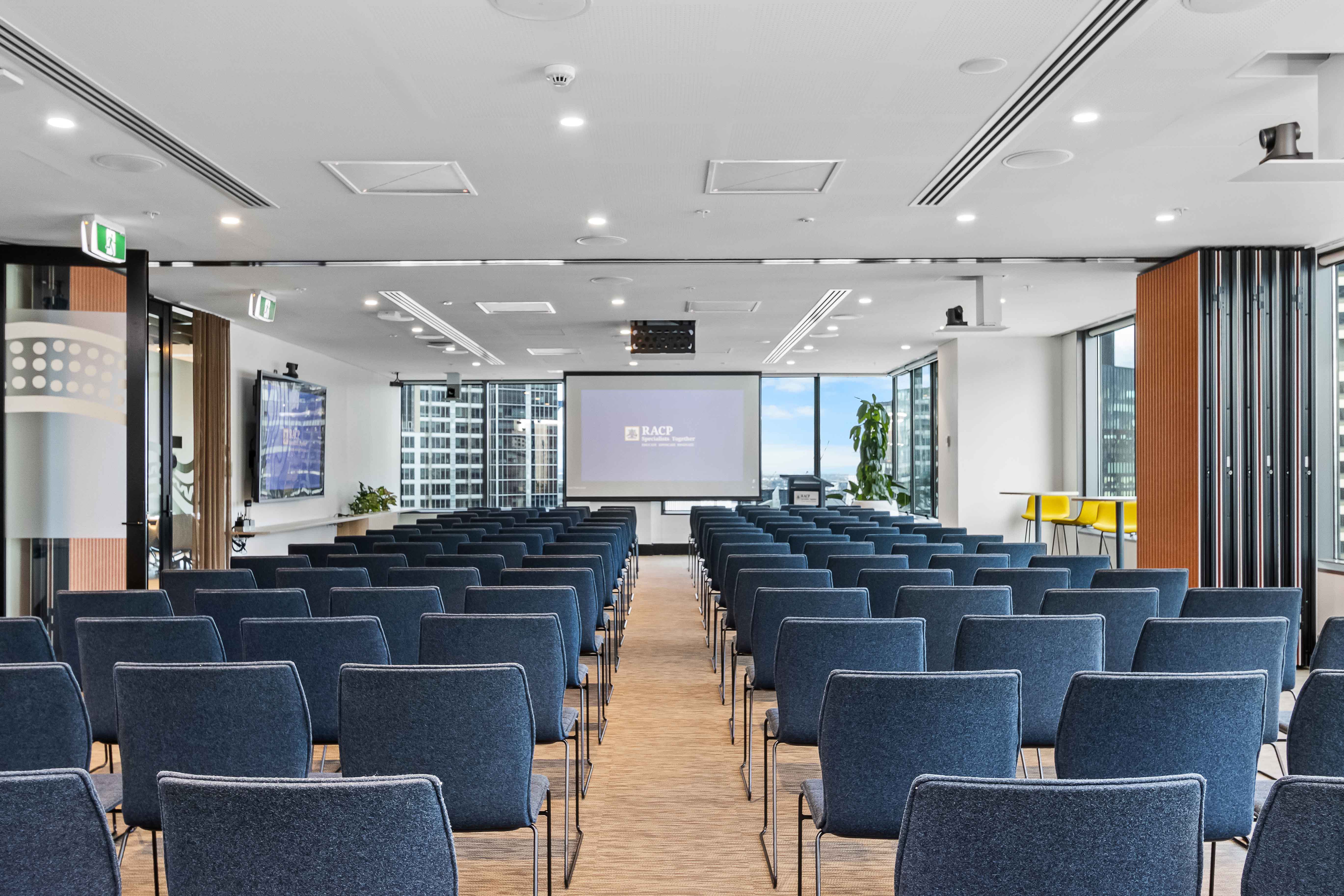 Modern conference room set up classroom-style with rows of blue chairs facing a projection screen and wall-mounted displays.