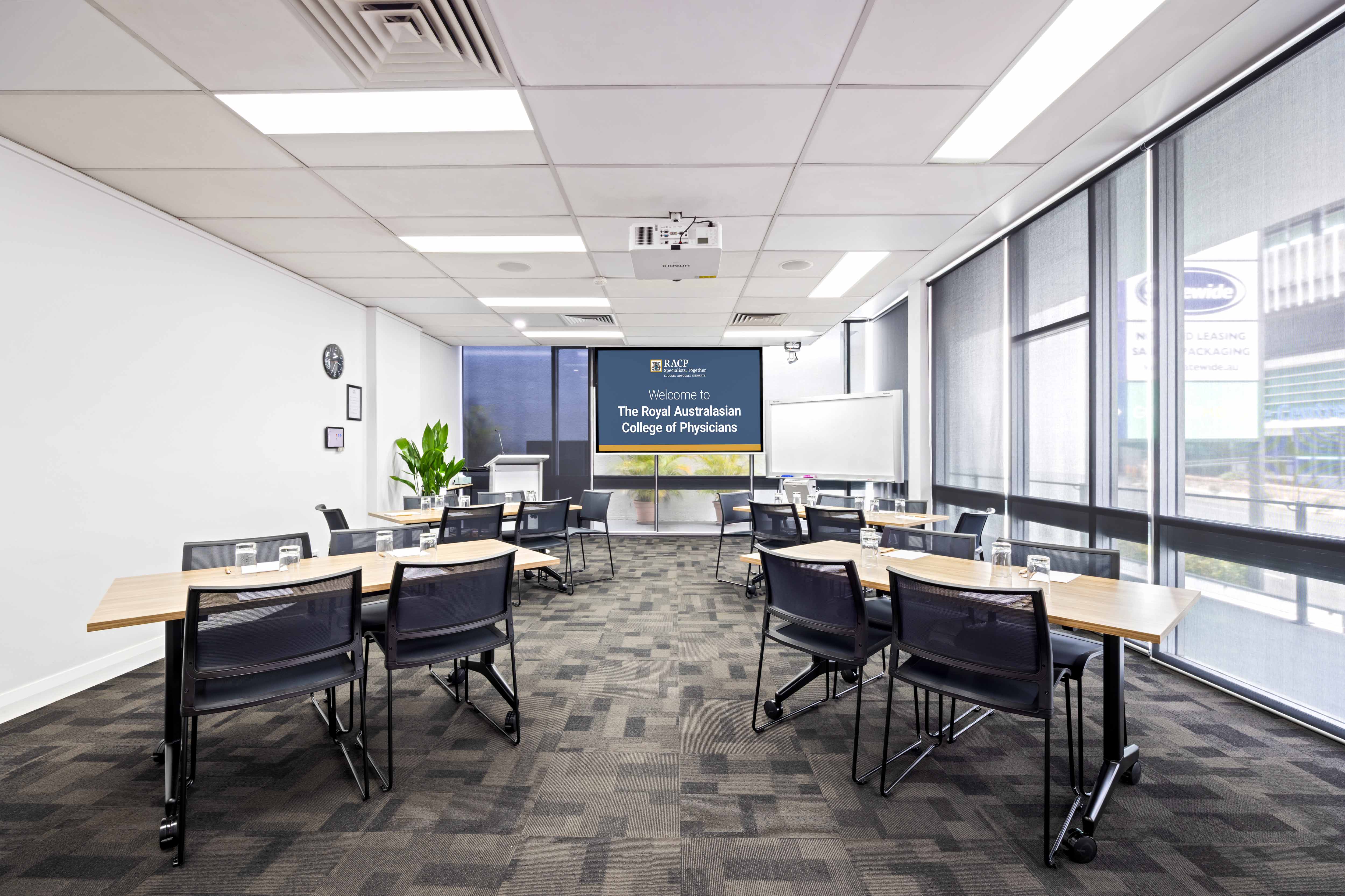 Meeting room with tables arranged in a classroom style facing a presentation screen displaying a Royal Australasian College of Physicians welcome slide.
