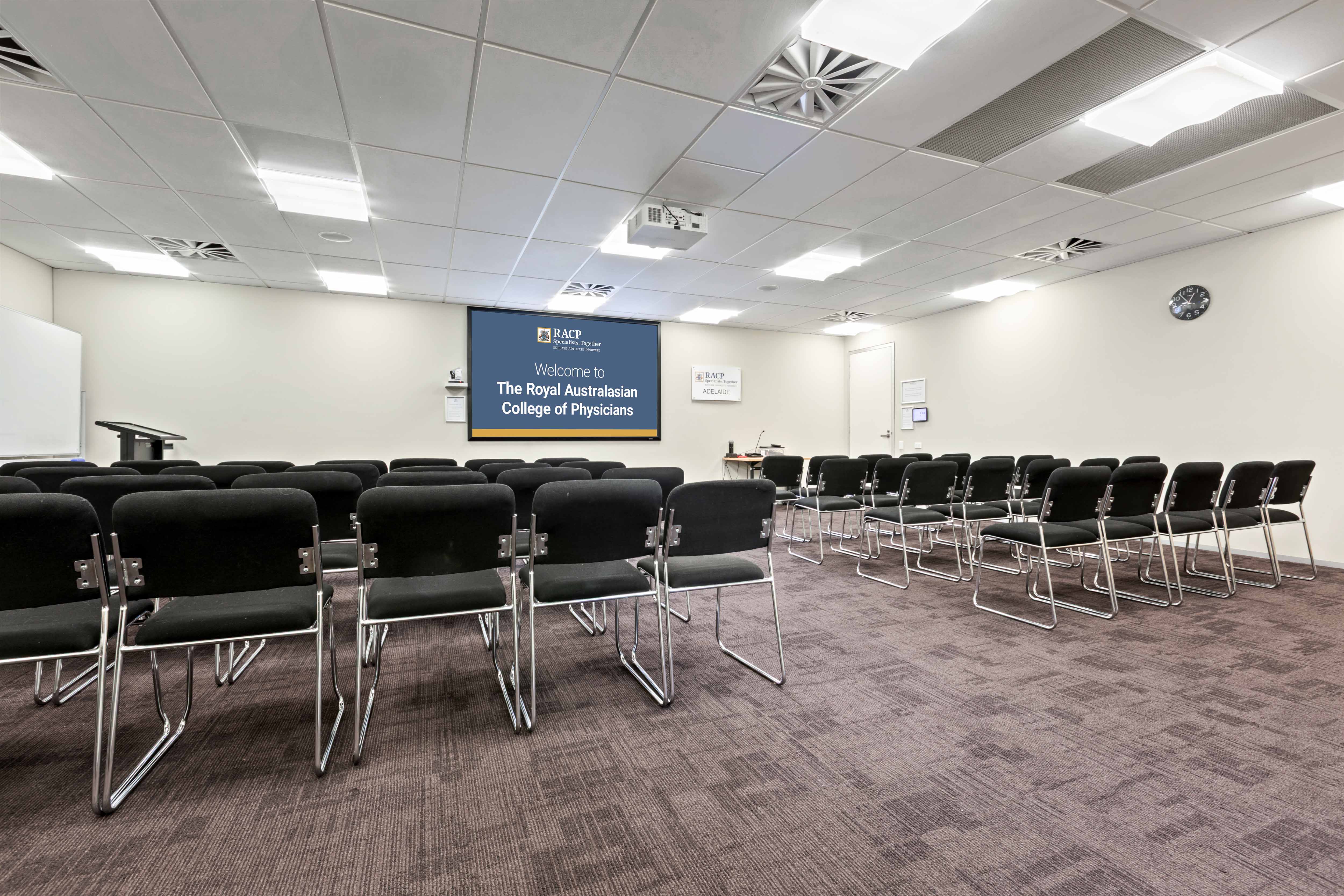 Conference room with rows of black chairs facing a presentation screen displaying a welcome message from the Royal Australasian College of Physicians.