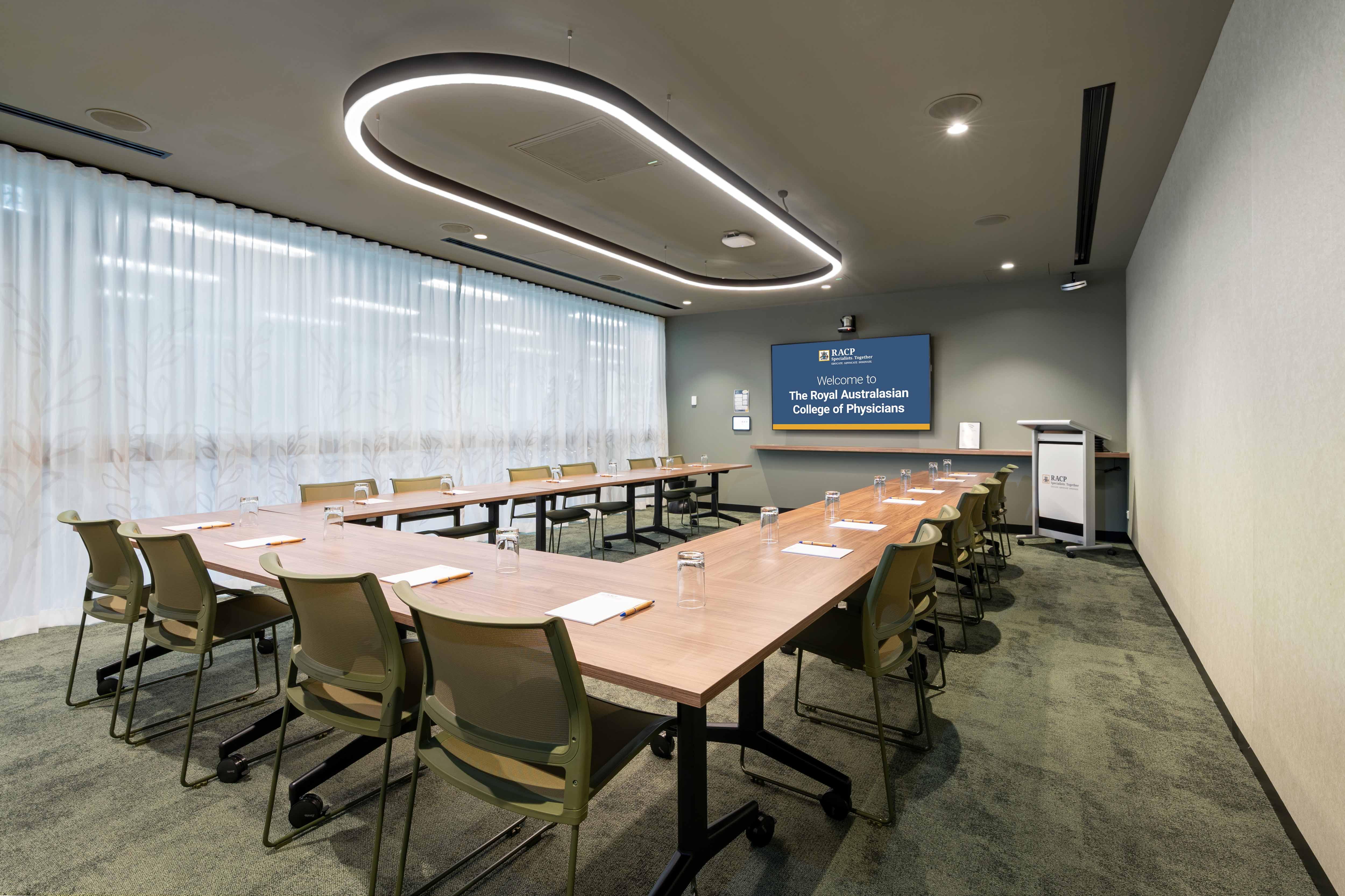 Meeting room with U-shaped tables and chairs facing a presentation screen displaying a Royal Australasian College of Physicians welcome slide.