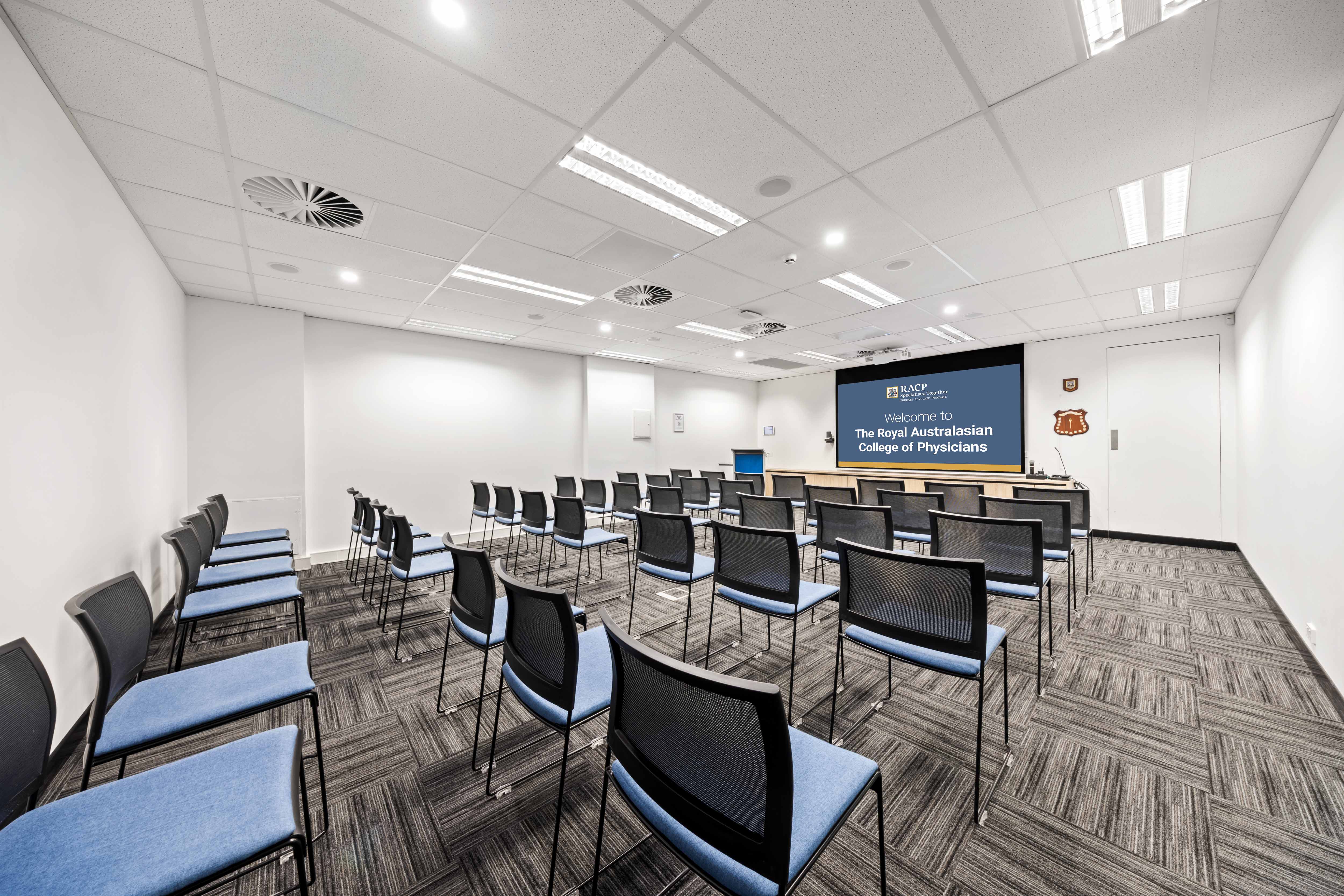Conference room with rows of chairs arranged in theatre style facing a presentation screen displaying a Royal Australasian College of Physicians welcome slide.