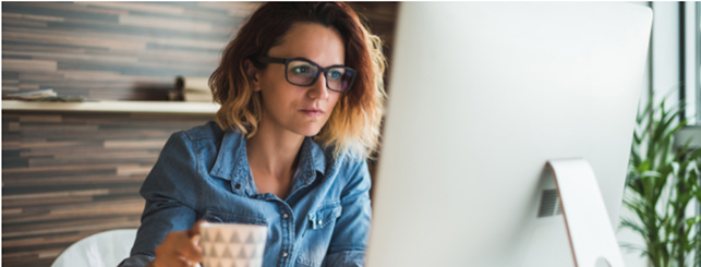 Woman in denim shirt holding a mug at a desk with a large monitor.