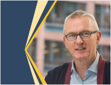 A man wearing a light blue shirt and dark cardigan stands in front of a building, with a graphic design element featuring dark blue and yellow angled shapes on the left side of the image.